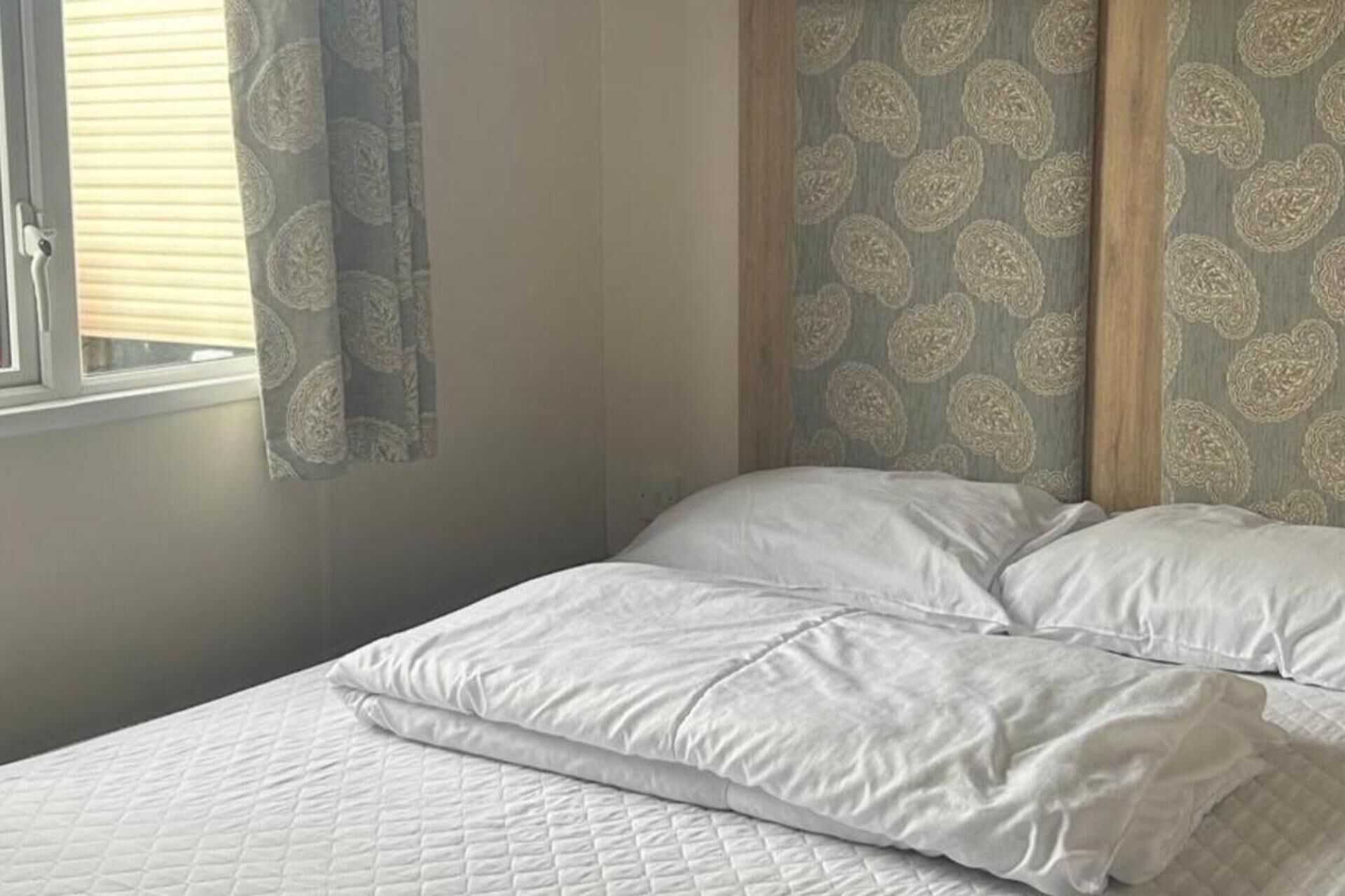A bedroom with a bed, white bedding, a window with blinds, and patterned wallpaper.