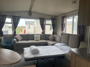 A cozy room with a sofa, a small table, and a bed with a rolled-up blanket. - Familycaravan-seaview-garden-ensuite-nearbeach (Clacton-on-Sea)