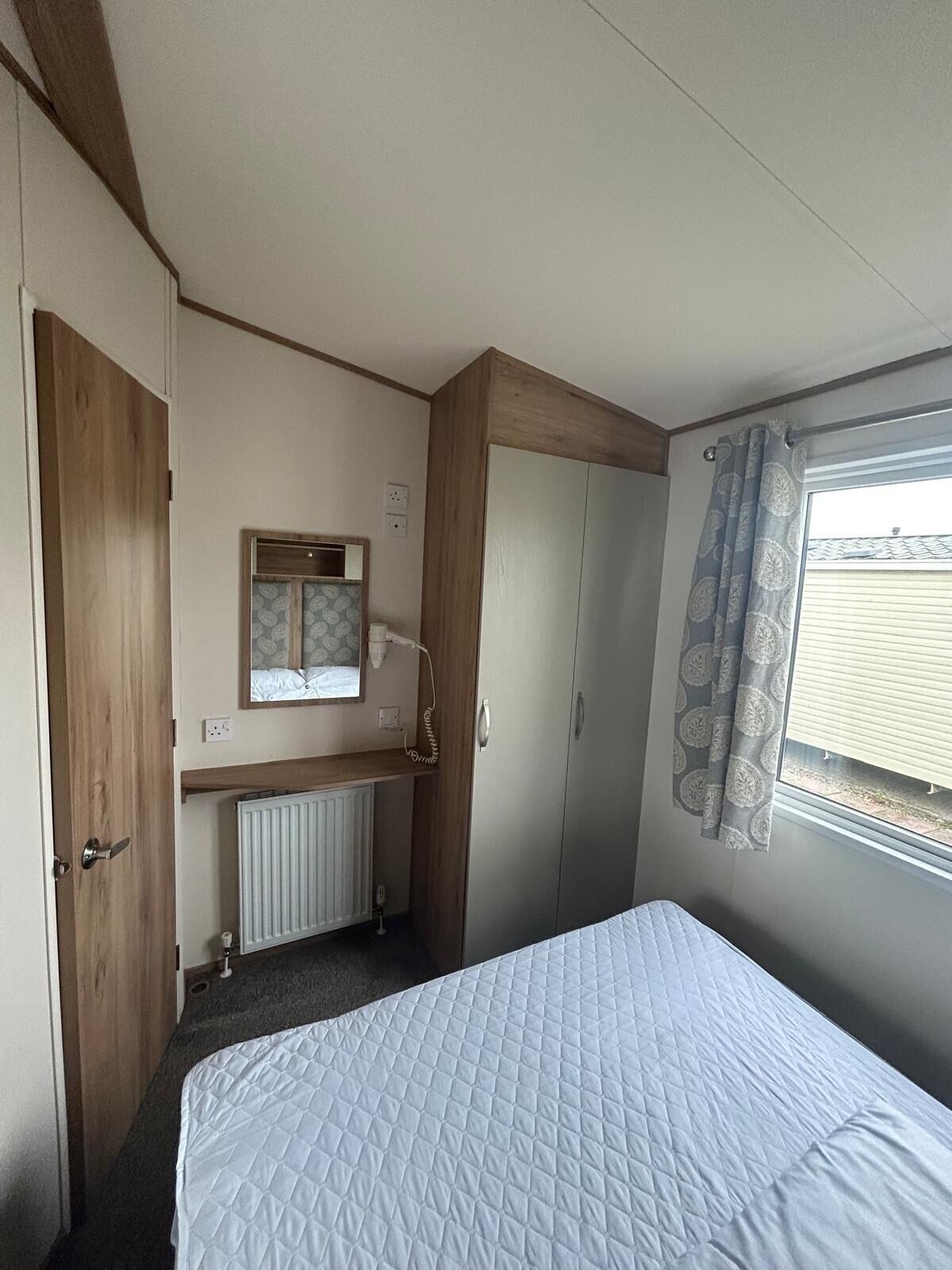 A compact room with a bed, a wardrobe, a small desk with a mirror, and a window with curtains.