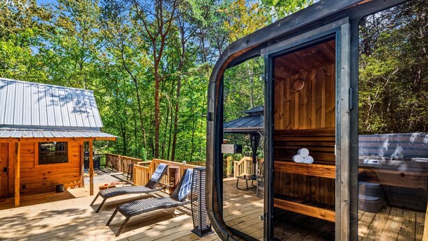 A wooden cabin with a deck, two lounge chairs, and a glass door leading to an interior space with a wooden bench and a table. - Intimate Cabin Escape: Hot Tub, Sauna, Firepit, Grill & Projector (Mentone)