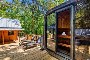 A wooden cabin with a deck, two lounge chairs, and a glass door leading to an interior space with a wooden bench and a table. - Intimate Cabin Escape: Hot Tub, Sauna, Firepit, Grill & Projector (Mentone)