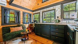 A cozy cabin interior with a wooden ceiling, a green sofa, a kitchen with dark cabinets, and large windows with a forest view. - Intimate Cabin Escape: Hot Tub, Sauna, Firepit, Grill & Projector (Mentone)