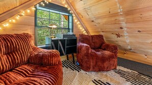 A cozy attic bedroom with a wooden ceiling, a large window with a view of greenery, a black entertainment unit, and two red velvet chairs. - Intimate Cabin Escape: Hot Tub, Sauna, Firepit, Grill & Projector (Mentone)