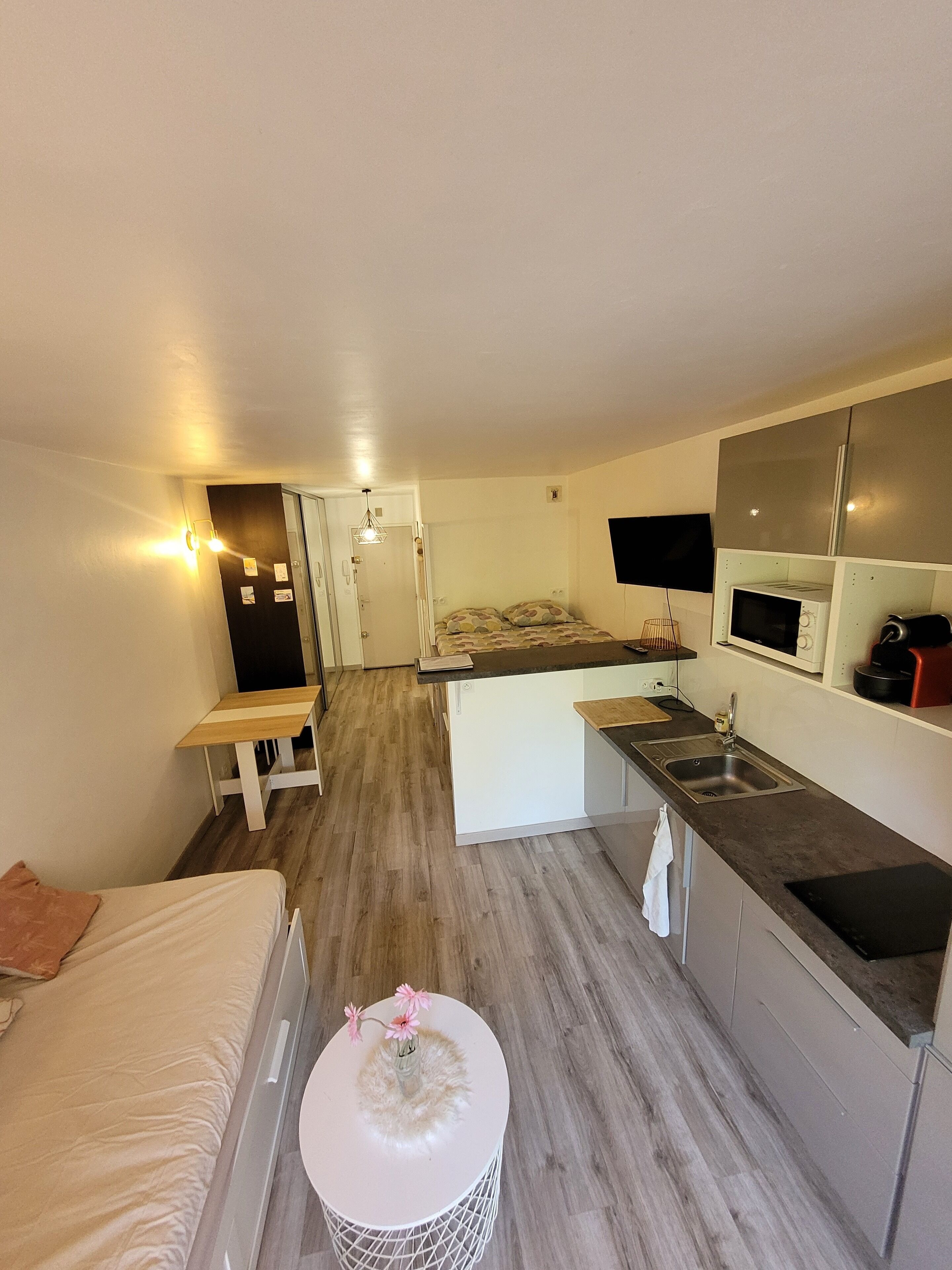 Een compacte hotelkamer met een bed, een klein rond tafeltje met een bloevas, een kitchenette met een magnetron en kookplaat, en een afzonderlijk slaapgedeelte met een slaapbank.