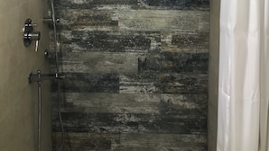 A shower with a rustic wooden wall design, a metal showerhead, and a white curtain. - Tatvamasi Homestay (New Delhi)