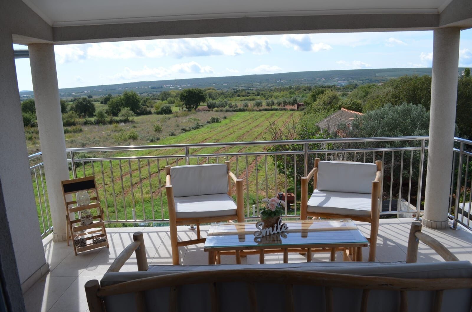 A balcony with two wooden chairs and a small table, overlooking a rural landscape with fields and a distant building.