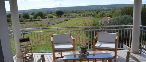 A balcony with two wooden chairs and a small table, overlooking a rural landscape with fields and a distant building.