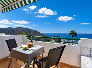 A balcony with a table set for two, overlooking a coastal landscape with mountains and the sea.