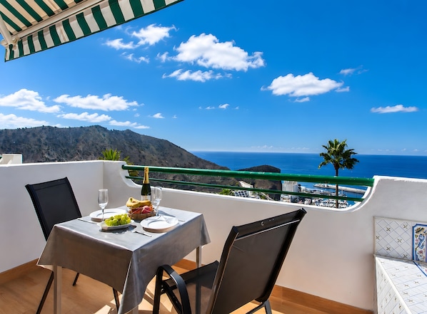 Un balcon avec une table dressée pour deux personnes, offrant une vue sur un paysage côtier avec des montagnes et la mer.