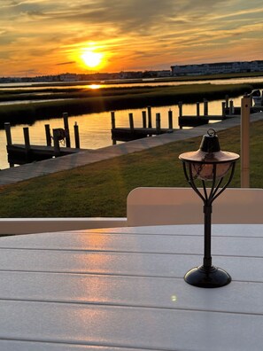 Une table avec une lampe sur un balcon donnant sur un coucher de soleil au-dessus d’une étendue d’eau, avec une jetée et des bâtiments en arrière-plan.