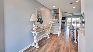 Un pasillo amplio con suelo de madera, una consola blanca con lámpara y una escalera que lleva a un balcón con mobiliario exterior.