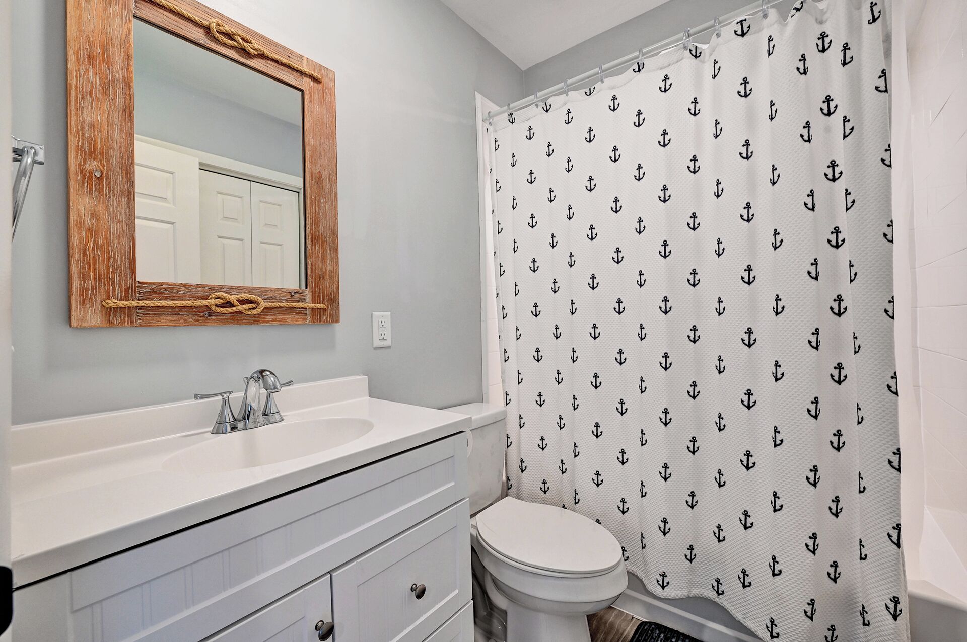 Un baño con espejo de marco de madera, lavamanos blanco, inodoro y cortina de ducha con motivos de anclas.