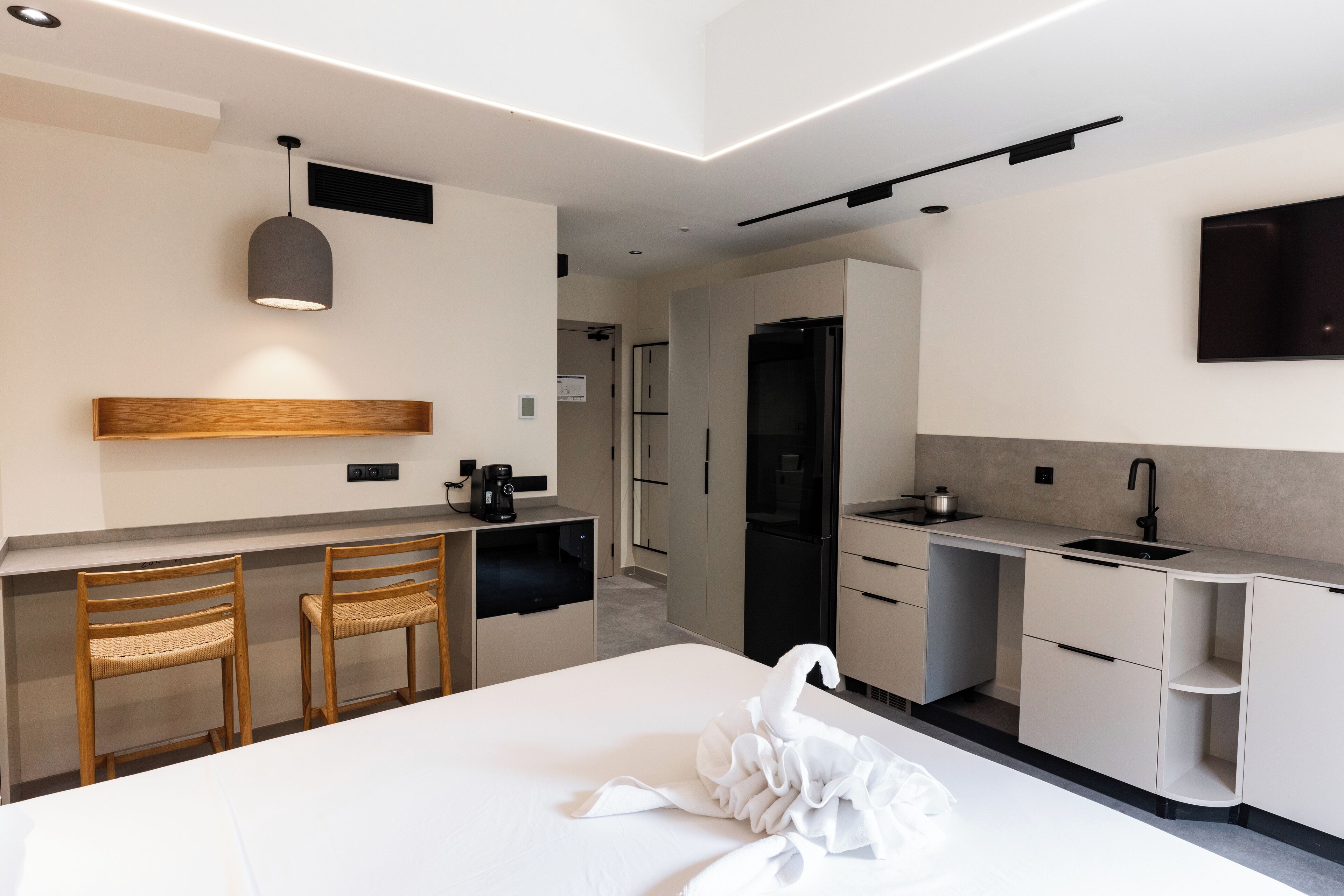 A modern hotel room with a large bed, a kitchenette, two wooden chairs, a white swan decoration, and a flat-screen TV mounted on the wall.