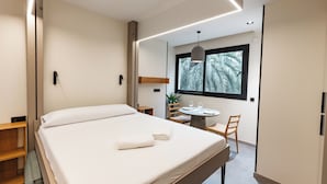 Une chambre d’hôtel moderne avec un grand lit, une petite table avec deux chaises, une fenêtre donnant sur des palmiers et un luminaire suspendu.