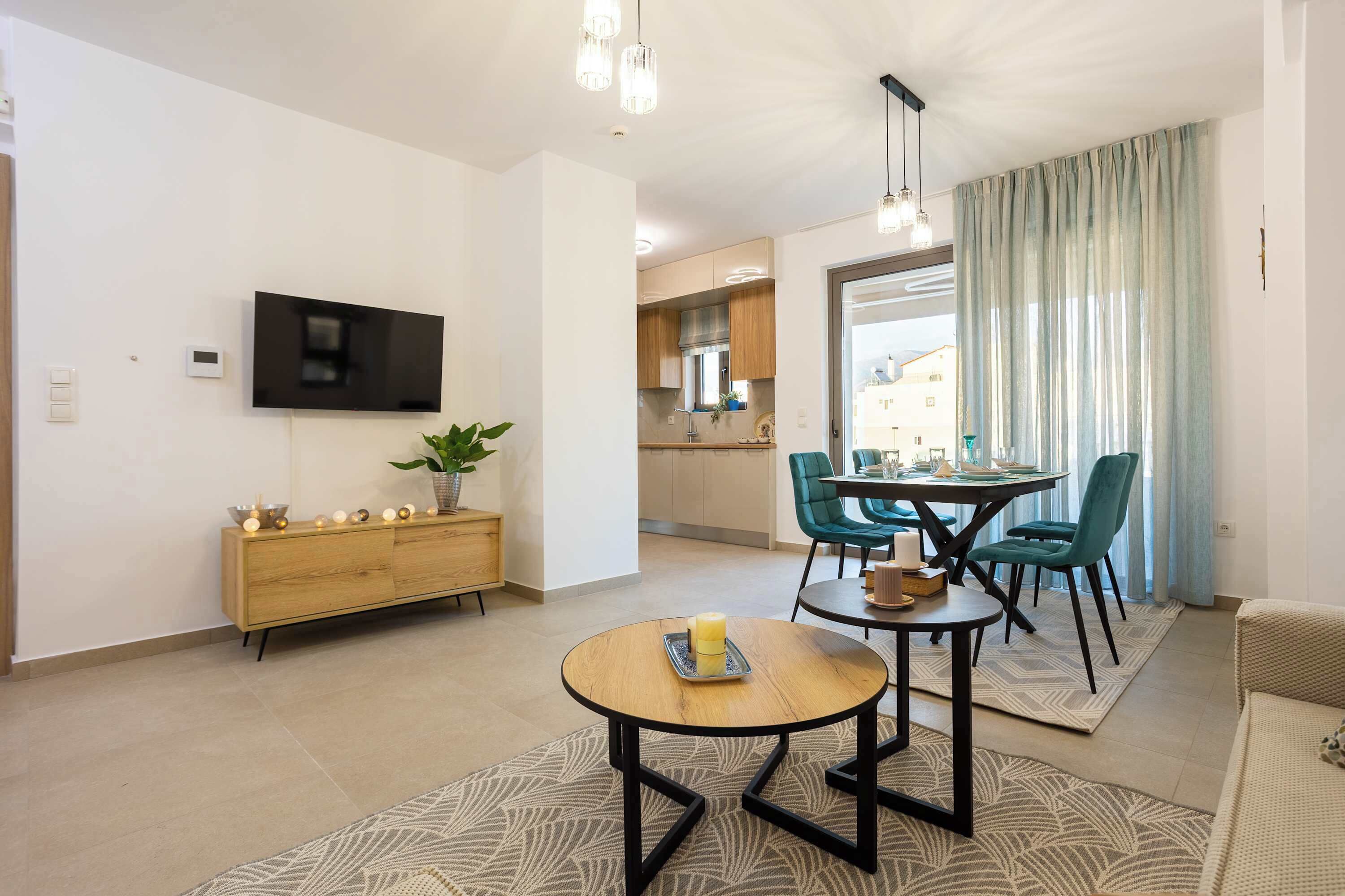 Uma sala de estar moderna com uma televisão de tela plana, um armário de madeira, uma mesa de jantar redonda com cadeiras e uma cozinha ao fundo.