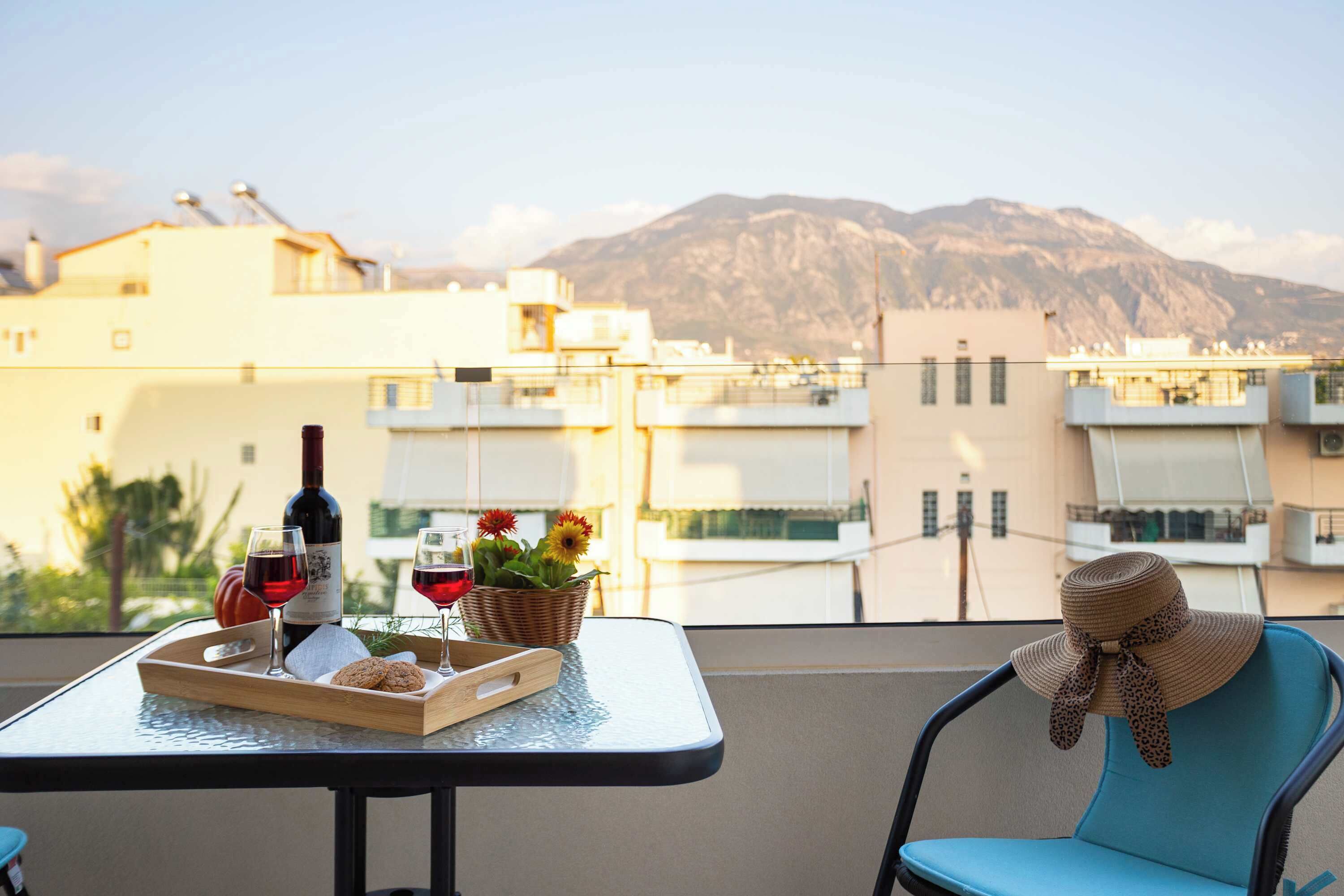 A balcony with a table set for two, featuring a bottle of wine, glasses, cookies, and a basket of flowers. A chair with a sun hat is also visible.