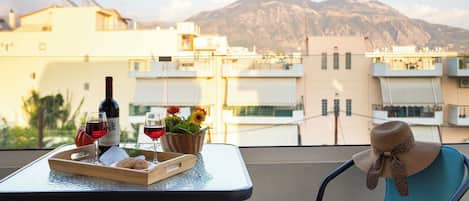 A balcony with a table set for two, featuring a bottle of wine, glasses, cookies, and a basket of flowers. A chair with a sun hat is also visible.
