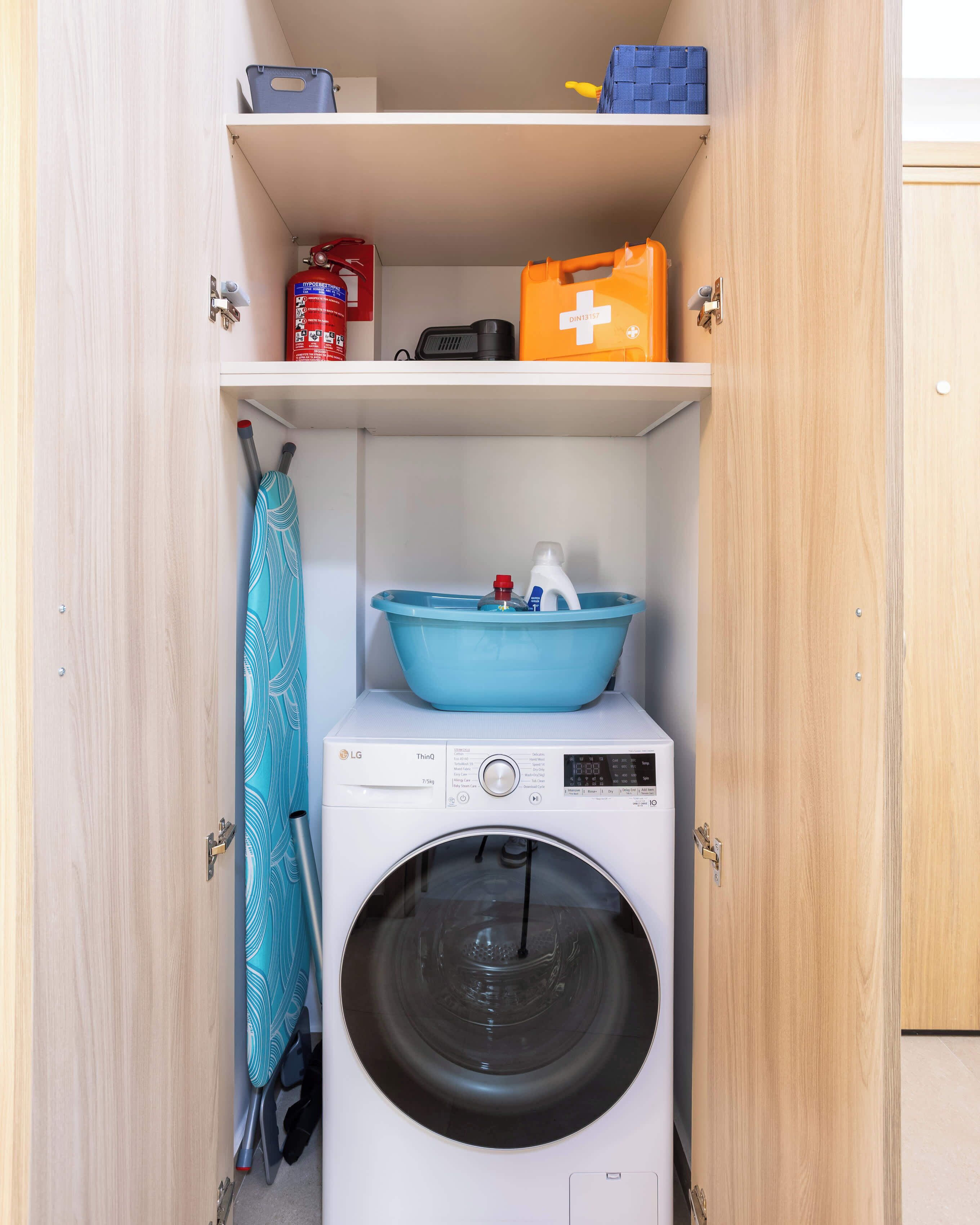 A compact laundry room with a front-loading washing machine, a blue basin on the top shelf, and a fire extinguisher on the left.