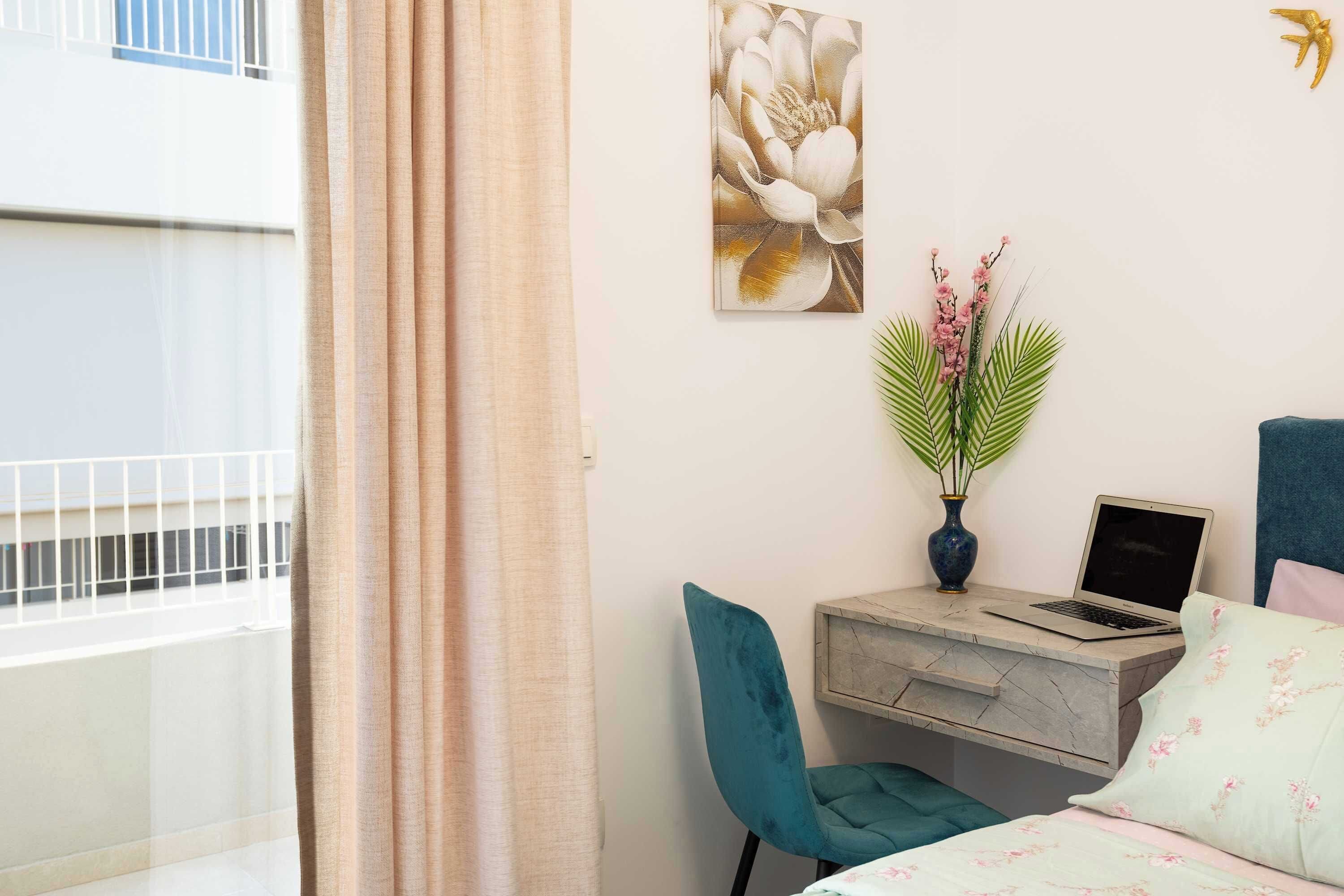 A bedroom with a bed, a desk with a laptop, a chair, a wall-mounted flower painting, and a window with curtains.