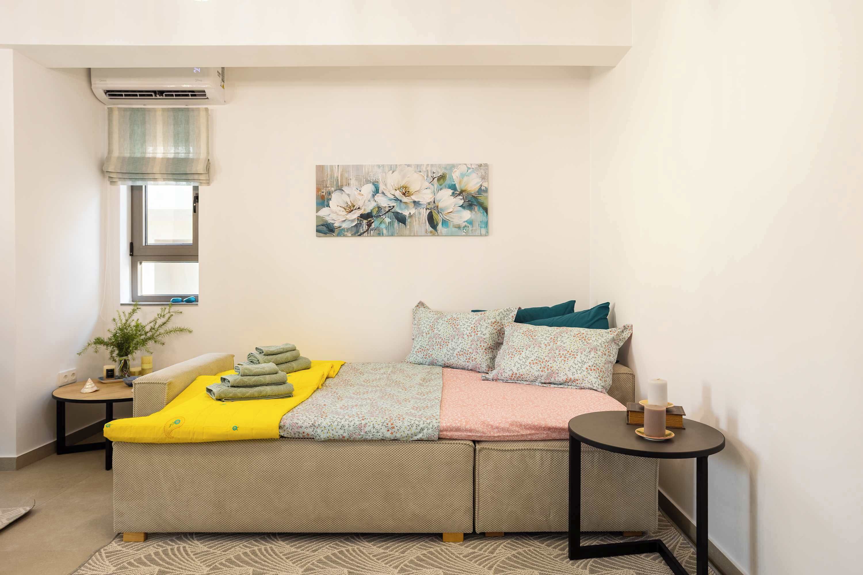A cozy bedroom with a sofa bed, a small bedside table, a potted plant, and a painting on the wall.