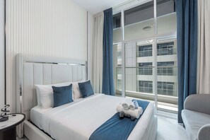 A hotel room with a large bed, a bedside table, a chair, and a view of a building through the window. - Al Barsha Studio Sleeps 2 in a Great Location (Dubai)