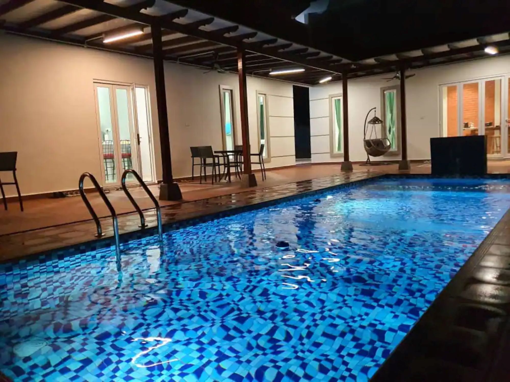 An indoor swimming pool with a mosaic tile floor, wooden deck, and a seating area with chairs and a hanging chair.