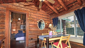 A cozy cabin interior with a wooden dining table, chairs, a bed, and a large window with curtains.