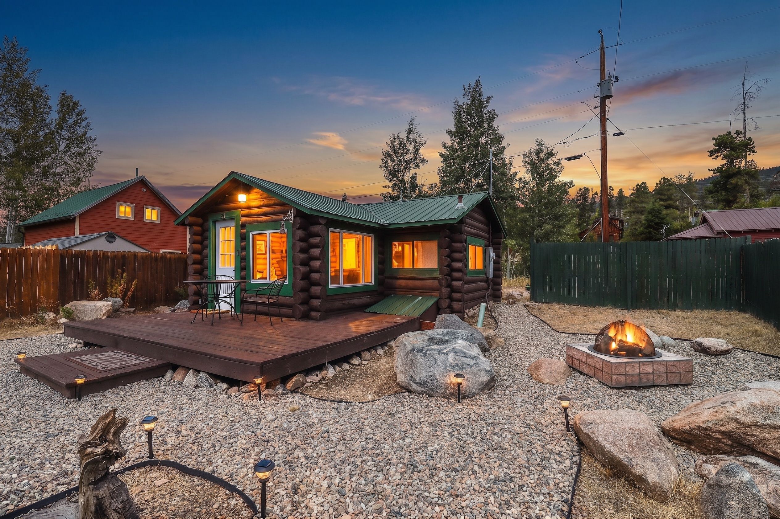 Une cabane en rondins dotée d’une terrasse et d’un foyer extérieur, entourée d’une zone recouverte de gravier et d’une clôture en bois.