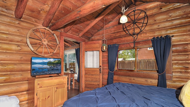 A log cabin room with a bed, wooden dresser, TV, and a large wheel hanging from the ceiling.