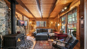 A cozy cabin interior with a stone wall, a wood-burning stove, a sofa, and a large window. - Classic Cabin in the Pines by Tahoe Getaways - Walk to Lake, Pet-Friendly 3 BR w/ Yard & Hot Tub! (Homewood)
