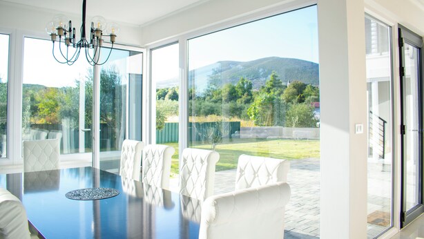 Un comedor moderno con una mesa grande y sillas blancas tapizadas, amplias ventanas que ofrecen vistas a un jardín ajardinado y colinas.