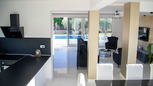 Una cocina moderna con encimera negra, estufa integrada y un refrigerador pequeño. Hay una puerta de vidrio que da a un patio con piscina y mobiliario exterior.