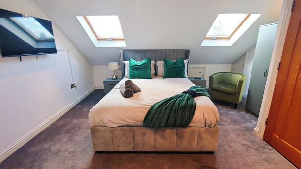 A bedroom with a large bed, two skylights, a TV mounted on the wall, a bedside table with a lamp, and a green armchair. - 6 en-suite Rooms all king beds  (Ilford)