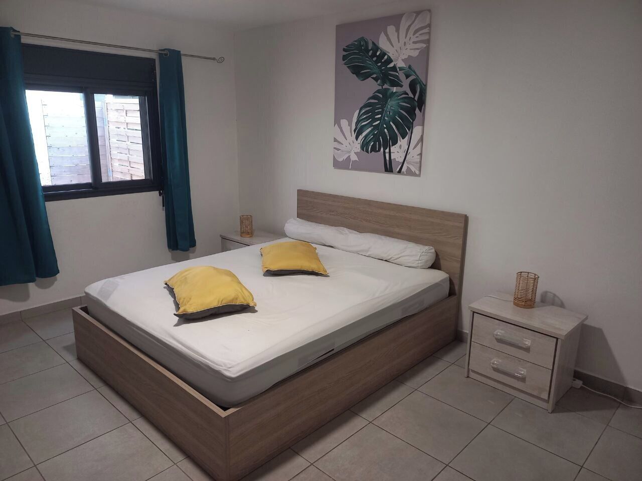 A bedroom with a bed, two pillows, a nightstand, a wall-mounted plant, and a window with curtains.