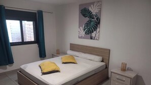 A bedroom with a bed, two pillows, a nightstand, a wall-mounted plant, and a window with curtains. - Comfortable house in a quiet area close to the sea and amenities in a seaside resort. (L'Étang-Salé les bains)