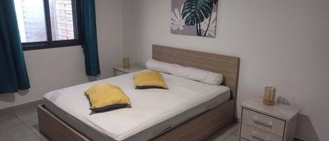 A bedroom with a bed, two pillows, a nightstand, a wall-mounted plant, and a window with curtains.