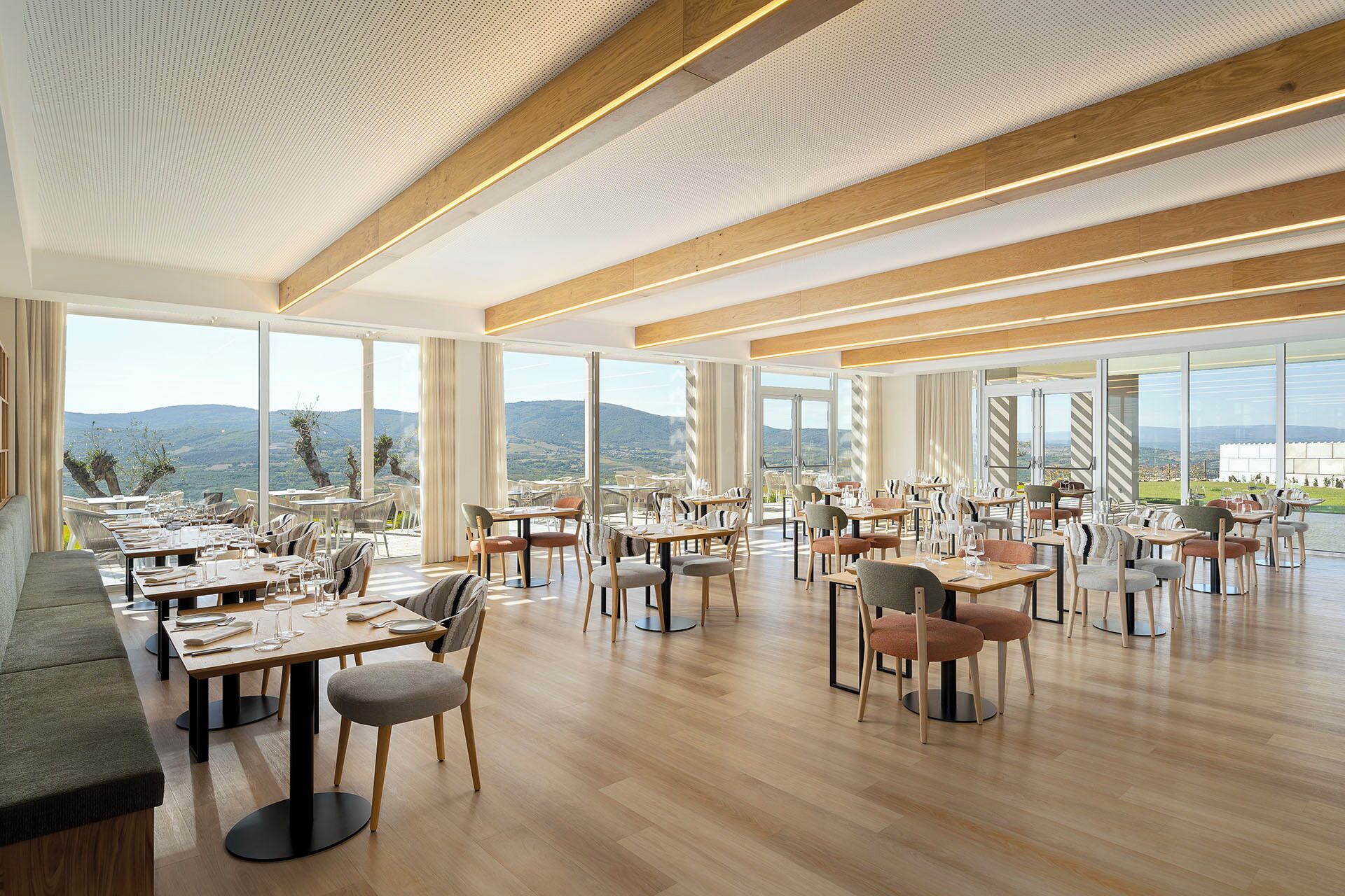 A spacious dining area with multiple tables and chairs, large windows offering a view of the landscape, and a wooden ceiling with integrated lighting.