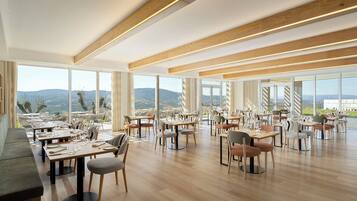 A spacious dining area with multiple tables and chairs, large windows offering a view of the landscape, and a wooden ceiling with integrated lighting.