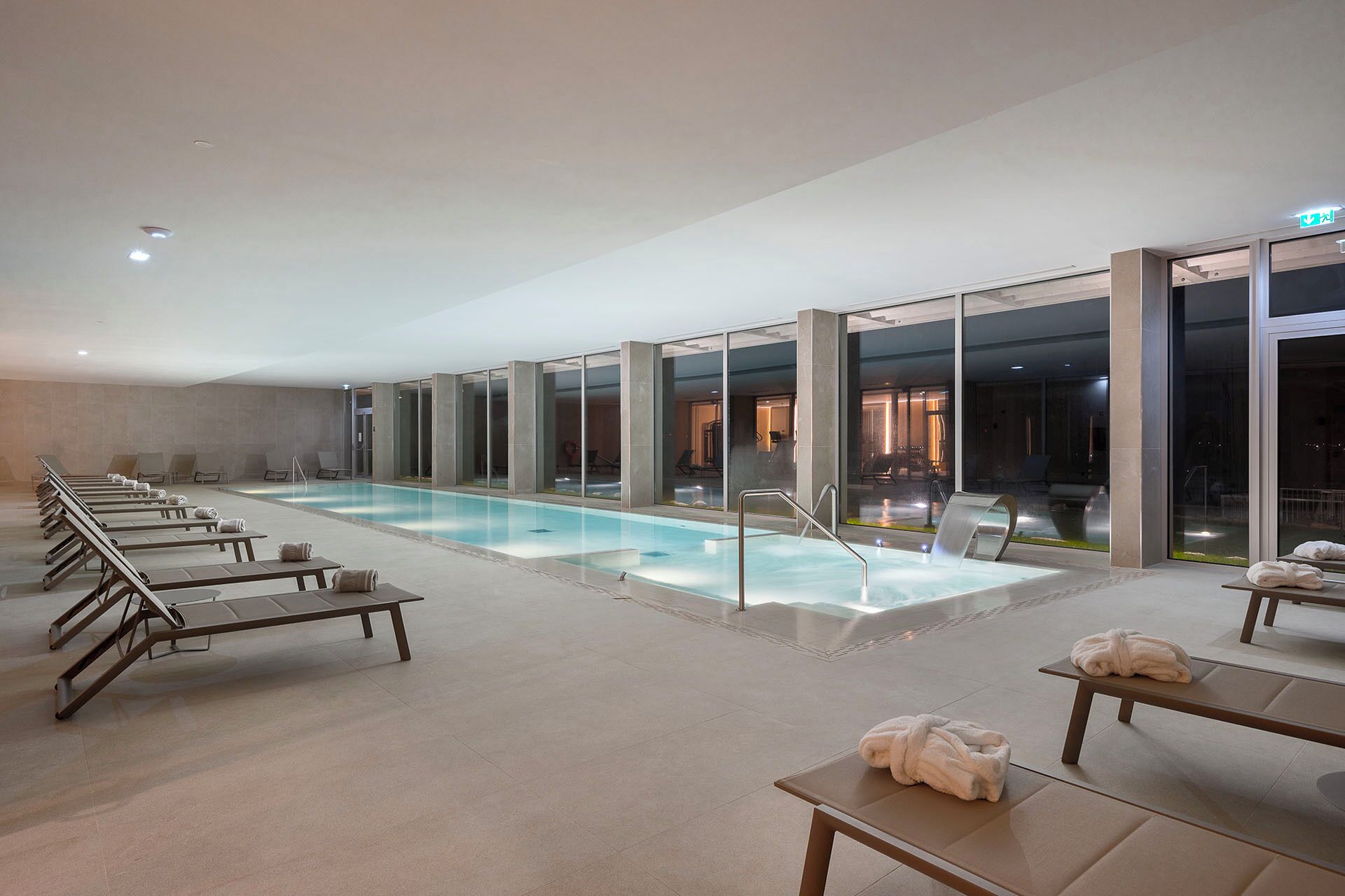 A modern indoor swimming pool with lounge chairs and a glass wall.