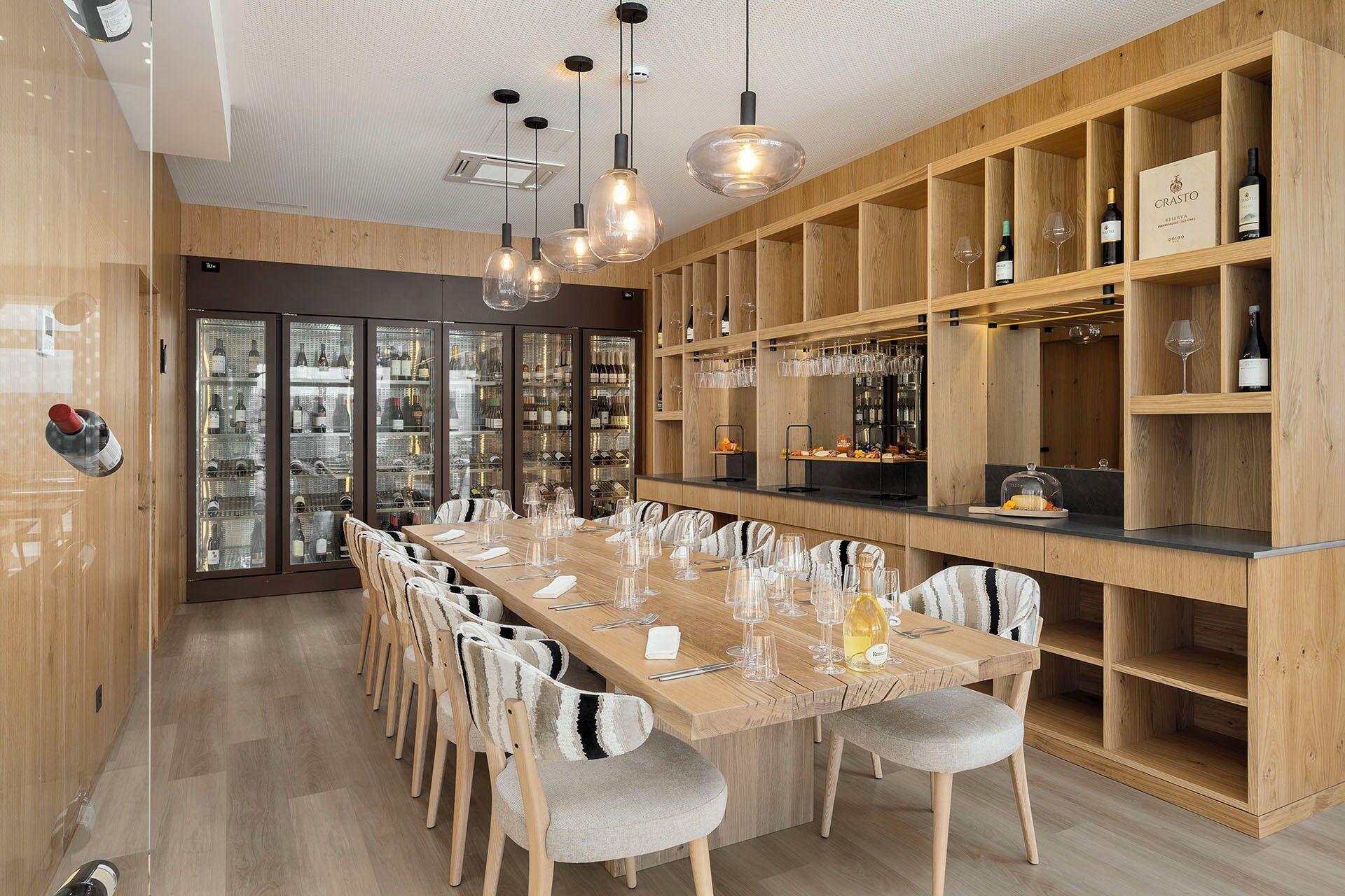 A restaurant with a long wooden table, chairs, and wine glasses. There are shelves with bottles and glasses on the right.