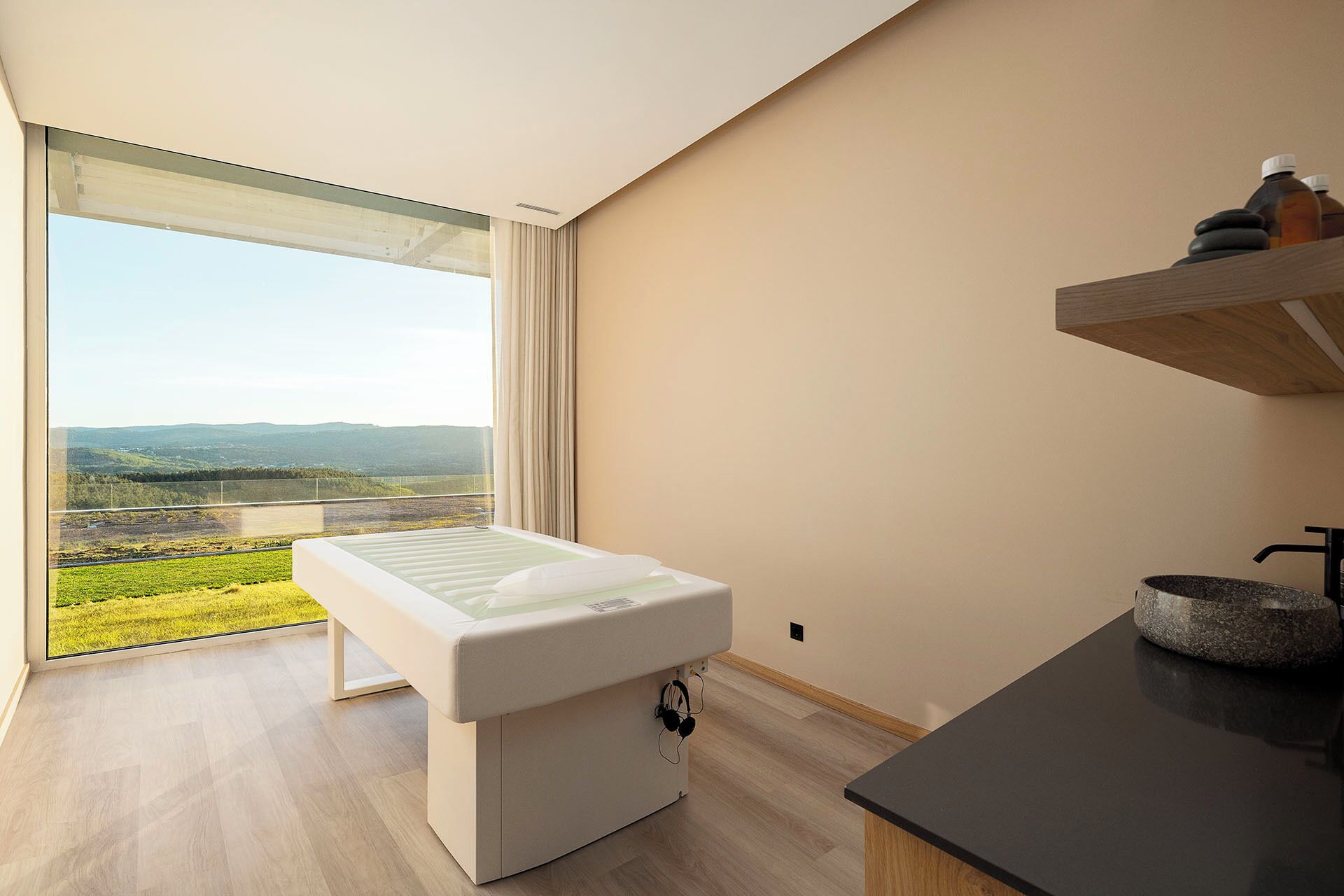Una habitación moderna con un ventanal grande, una camilla de masaje, un estante de madera con botellas y una encimera negra.