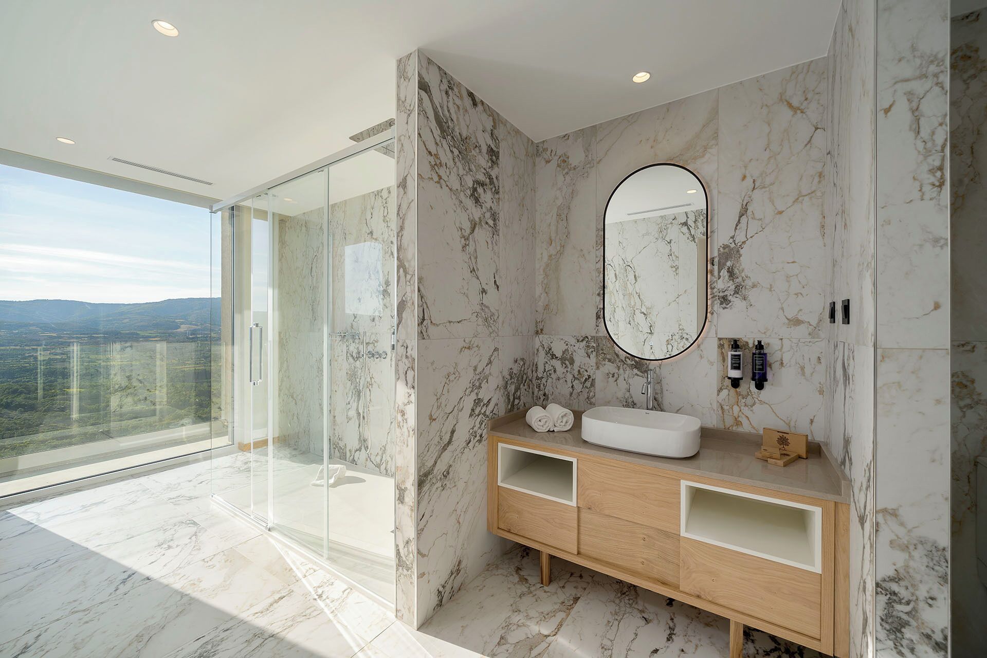 Une salle de bain moderne avec un mur en marbre, un grand miroir, un meuble-lavabo en bois et une cabine de douche en verre.