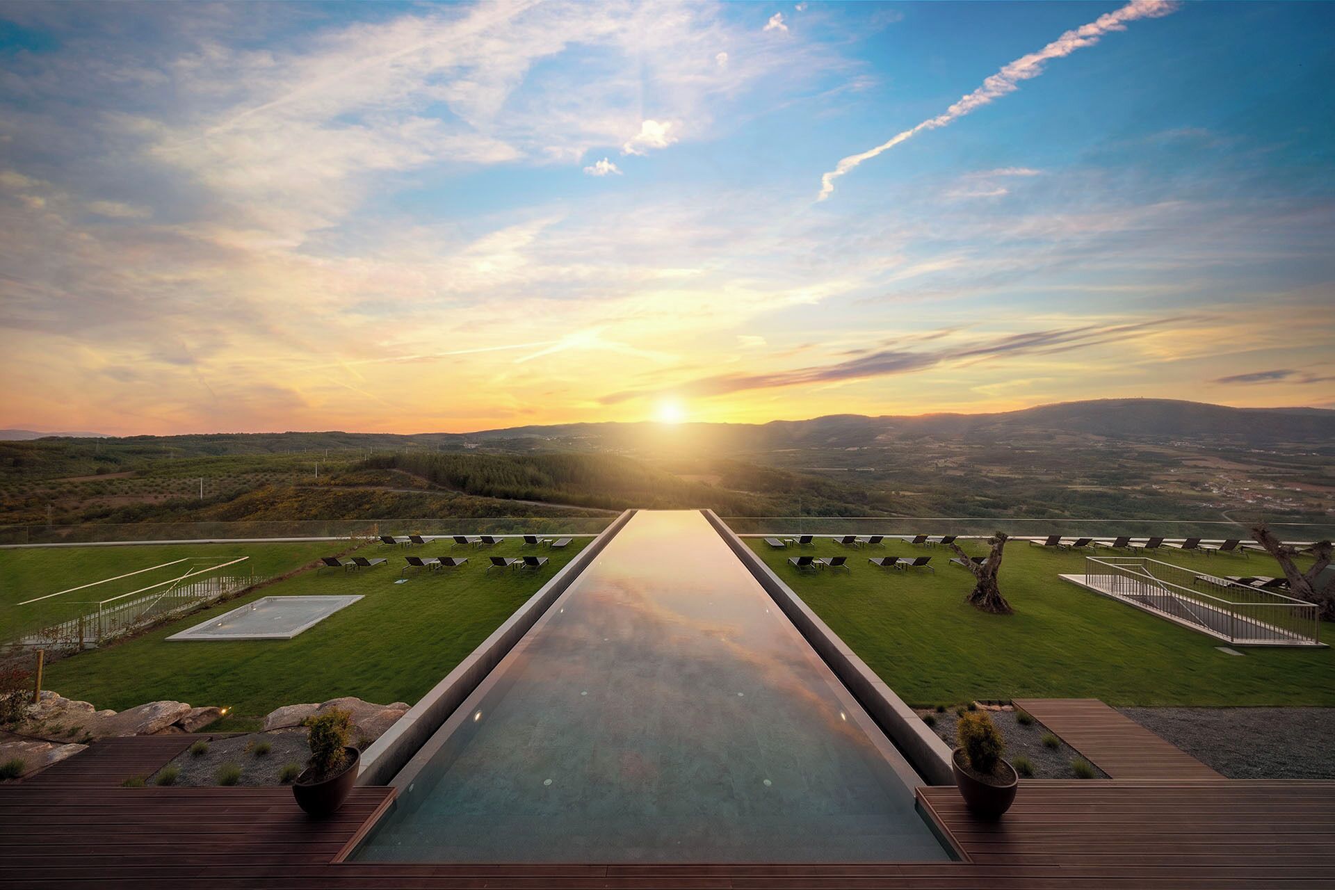 A long, reflective water feature leading to a deck with a scenic view of a valley and mountains during sunset.