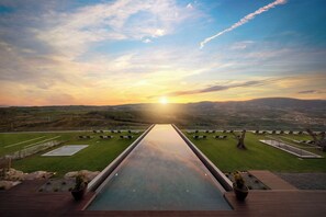 Un long bassin d’eau décoratif, invitant à la contemplation, mène à une terrasse offrant une vue panoramique sur une vallée et des montagnes au coucher du soleil.