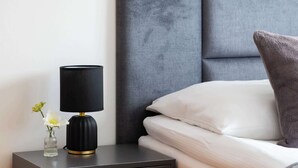 Una cama bien hecha con cabecera gris, una mesita de noche pequeña con una lámpara negra y un cajón de madera.