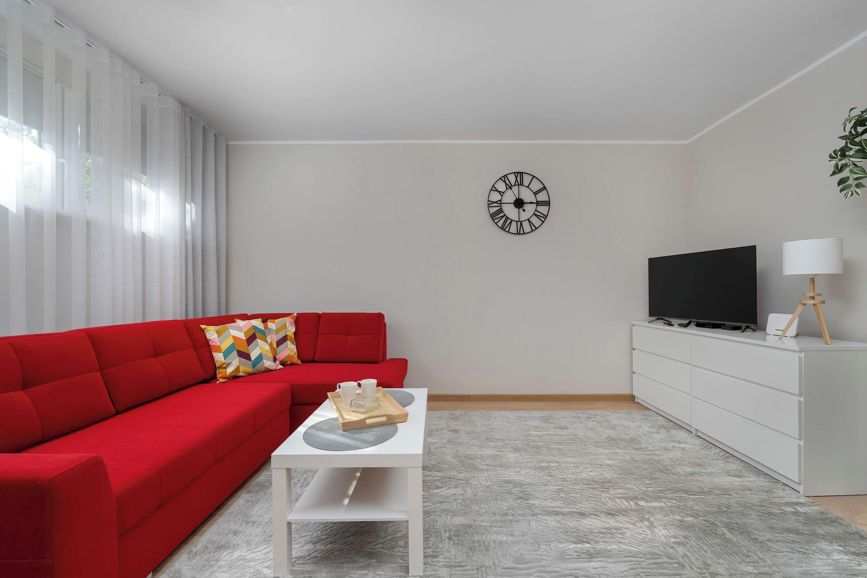 A living room with a red sofa, a white coffee table with a tray, a TV on a white cabinet, and a clock on the wall.