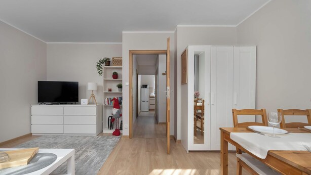 A modern living room with a TV, white furniture, a dining table, and a hallway leading to a kitchen. - Intimate Apartment | Two Bedrooms, Work Desk (Wrocław)