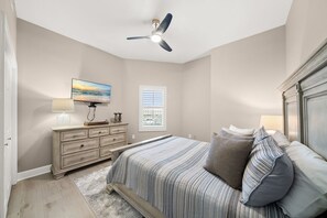 Een slaapkamer met een bed, een dressoir, een plafondventilator, een schilderij en een raam met jaloezieën.