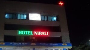 A hotel named "HOTEL NIRALI" with a signboard, a bank named "Indian Bank" below it, and various shops at the ground level. - Hotel Nirali Rooms - Nexus Hospitality (Surat)
