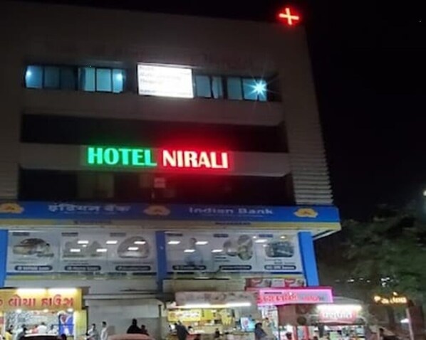 A hotel named "HOTEL NIRALI" with a signboard, a bank named "Indian Bank" below it, and various shops at the ground level. - Hotel Nirali Rooms - Nexus Hospitality (Surat)